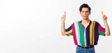 Şüpheci ve hoşnutsuz genç adam parmaklarını havaya kaldırıyor, hayal kırıklığına uğruyor, beyaz arka plana karşı duruyor..