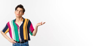 Kafası karışmış eşcinsel adam garip bir şeye bakıyor, elini kaldırıyor habersiz ve şaşkın, beyaz arka planda duruyor..