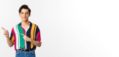 Genç, yakışıklı, eşcinsel bir adam logoya bakıyor, gülümsüyor, beyaz arka planda duruyor..