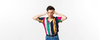 Endişeli genç eşcinsel adam, panikle ayakta duruyor, ellerini başının üstüne koyuyor ve sorunlu bir şekilde aşağı bakıyor, beyaz arka planda duruyor..