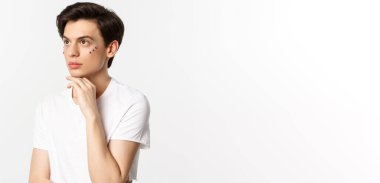 Millet, lgbtq ve güzellik konsepti. Güzel çift cinsiyetli adam, gözlerinin altında pırıltı, sol üst köşeye bakıyor, beyaz arka plana karşı poz veriyor..