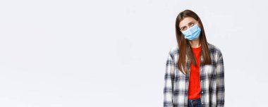 Coronavirus salgını, karantina, sosyal mesafeler ve duygu kavramı. Hüzünlü ve sıkkın, tıbbi maskeli, üzgün genç bir kadın, iç çekerek başka tarafa bakıyor, bıkkın bir şekilde evde oturuyor..