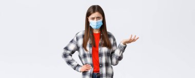 Coronavirus salgını, karantina, sosyal mesafeler ve duygu kavramı. Kafası karışmış ve canı sıkkın genç bir kadın bunu anlayamaz, omuz silker, şaşkın kamera, tıbbi maske.