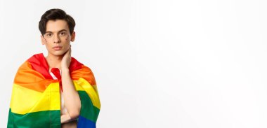 Yüzünde sim olan güzel bir gay adamın belden yukarı pozu, kendini gökkuşağı lgbt bayrağıyla sar ve yüzüne nazikçe dokun, kameraya bak, beyaz arkaplan.