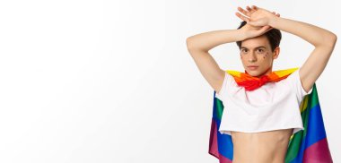 Yüzünde parıltı olan güzel bir gay adam, açık bluz ve gökkuşağı lgbt bayrağı giyiyor, beyaz arka planda poz veriyor..