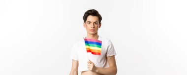 Gurur ve lgbtq kavramı. Gökkuşağı bayrağı tutan, yüzünde parıltı olan ve beyaz arka planda duran çekici ve kadınsı bir adamın belden yukarı pozu..