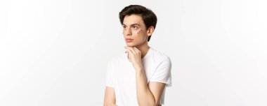 Millet, lgbtq ve güzellik konsepti. Güzel çift cinsiyetli adam, gözlerinin altında pırıltı, sol üst köşeye bakıyor, beyaz arka plana karşı poz veriyor..
