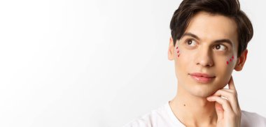 Millet, lgbtq ve güzellik konsepti. Uygulamalı dudak parlatıcısı ve pırıltısı olan mutlu eşcinsel adamın yakın çekimi, gülümsüyor ve sol üst köşeye bakıyor, beyaz arka planın üzerinde duruyor..