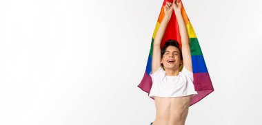 Gururlu ve mutlu gay adamın lgbtq gökkuşağı bayrağını dalgalandırması, rahatlamış duygularla gülümsemesi, kot pantolon giymiş, beyaz arka plan.