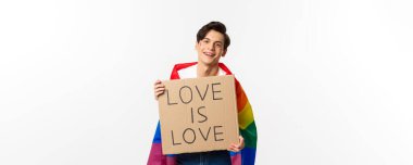 Gülümseyen eşcinsel eylemci elinde işaret işareti olan aşk, lgbt onur geçidine aşktır, Gökkuşağı bayrağı takar, beyaz arka planda durur..