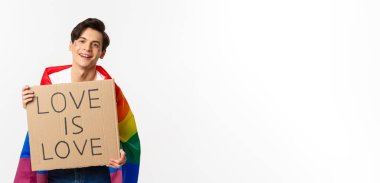 Gülümseyen eşcinsel eylemci elinde işaret işareti olan aşk, lgbt onur geçidine aşktır, Gökkuşağı bayrağı takar, beyaz arka planda durur..