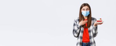 Coronavirus salgını, sosyal mesafeli yaşam tarzı ve tatil kutlamaları konsepti. Medikal maskeli çekici bir kadın elinde doğum günü pastası tutuyormuş gibi bir dilek tutuyor..