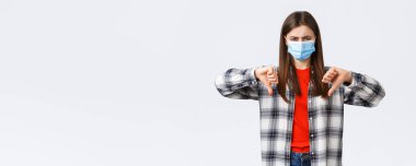 Coronavirus salgını, karantina, sosyal mesafeler ve duygu kavramı. Tıbbi maske takmış hayal kırıklığına uğramış ve kızgın genç bir kadın karşı oy kullandı, baş parmağını kaldırdı ve yüzünü buruşturdu.