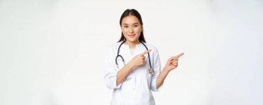Sağlık hizmeti, tıp insanları konsepti. Gülümseyen Asyalı kadın doktor, parmaklarını sağa gösteriyor, reklam veriyor, hastane bilgilerini gösteriyor, beyaz arka planda duruyor..