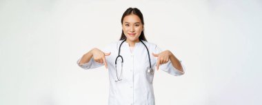 Genç Asyalı kadın doktor, parmaklarını eğip gülümsüyor, steteskop ve sağlık üniforması giyiyor, beyaz arka planda duruyor..