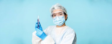 Asyalı hemşire ya da laboratuvar çalışanı kişisel koruma ekipmanında test tüpü gösteriyor, mavi arka planda tıbbi maske takıyor..