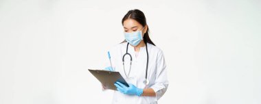 Hasta bilgilerini panoya yazan, reçete yazan ya da teşhis koyan Asyalı bir kadın doktor. Plastik eldivenli, beyaz arka planlı, tıbbi maske takıyor..