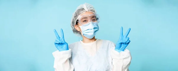 Happy asian female doctor, nurse in personal protective equipment, face mask and sterile gloves, smiling and showing peace, v-signs, blue background.