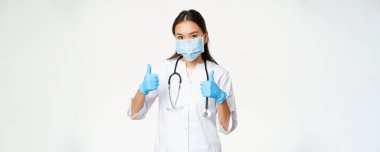 Covid-19 ve sağlık çalışanları konsepti. Yüzünde maske ve lastik eldiven olan gülümseyen Asyalı kadın doktor baş parmak gösteriyor, beyaz arkaplan.