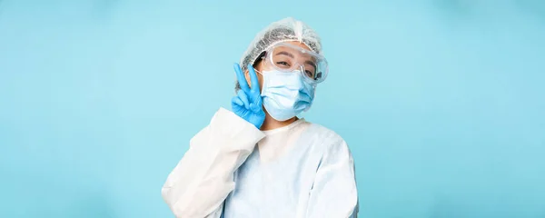 Happy, kawaii asian female doctor, nurse in personal protective equipment, showing v-sign peace gesture near face mask, blue background.