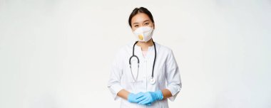 Solunum cihazında gülümseyen Asyalı kadın doktor, tıbbi maske ve lastik eldivenler, yardıma hazır, beyaz arka planda klinik üniforması içinde duruyor. Covid-19 ve karantina konsepti.