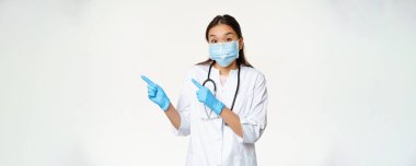 Heyecanlı Asyalı kadın doktor solu işaret ediyor, önleyici tedbir olarak tıbbi maske takıyor, beyaz arka planda üniforma giyiyor..