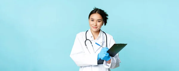 Smiling asian medical worker, female physician writing down patient info, holding pen and clipboard, standing in uniform over blue background.