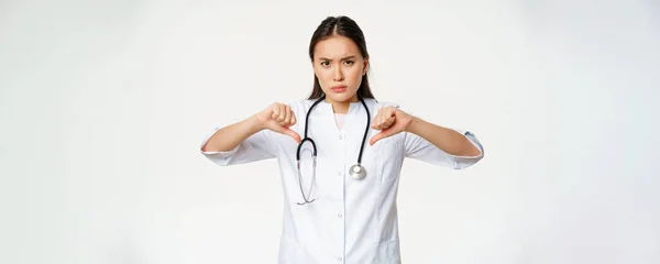 失望した深刻なアジアの女性医師 病院の医師は 親指を示しています不承認で 笑顔悪い嫌い 白い背景の上に立って — ストック写真