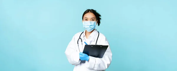 Covid-19 and quarantine concept. Smiling asian female physician, nurse in face mask holding clipboard, shift at hospital, standing over blue background.
