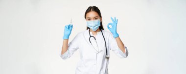 Covid-19 aşısı ve sağlık sigortası. Tıbbi maske ve eldiven takan Asyalı kadın doktor, elinde aşı şırıngası var ve iyi bir işaret, beyaz arka plan..