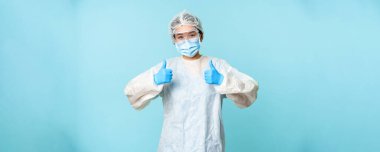 İyimser Asyalı hemşire ya da doktor, kişisel koruyucu ekipman giyiyor, baş parmaklarını gösteriyor, koronavirüs önleme kampanyası, mavi arka plan..