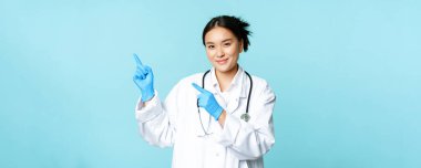 Gülümseyen kadın doktor, tıp üniformalı fizikçi ve steril eldiven giyen, tanıtım için sol parmakları gösteren, klinik logosu, mavi arka planda duran..