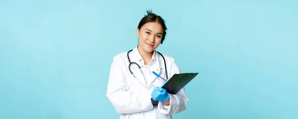 Portrait of female doctor, asian physician in medical uniform, writing down patient info, standing against blue background. Healthcare concept