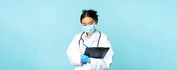 Image of korean doctor, female nurse in medical face mask, writing down patient information, examining on hospital shift, standing in uniform over blue background.