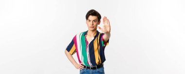 Genç kendine güvenen eşcinsel adam hayır diyor, dur işareti gösteriyor ve hoşnutsuz görünüyor, beyaz arka planda duruyor..