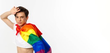 Yüzünde parıltı olan güzel bir gay adam, açık bluz ve gökkuşağı lgbt bayrağı giyiyor, beyaz arka planda poz veriyor..