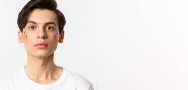 Millet, lgbtq ve güzellik konsepti. Yüzünde sim olan, beyaz arka planda duran, kameraya bakan, çift cinsiyetli güzel bir adamın yakın çekimi..