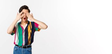 Yakışıklı, genç, eşcinsel bir adamın görüntüsü şaşkınlığı görmek için gözleri açık, parmaklarının arasından şaşkınlıkla bakan, beyaz arka planda duran.