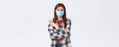 Coronavirus salgını, karantina, sosyal mesafeler ve duygu kavramı. Kafası karışmış ve endişeli bir kız, tuhaf bir soru soruyor, parmağıyla sol tarafı işaret ediyor, tıbbi maske takıyor..