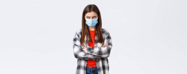 Coronavirus salgını, karantina, sosyal uzaklık ve duygu konsepti. Tıbbi maskeli, kolları çapraz göğüslü, alnının sol üst köşesinden bakan kıskanç ya da kızgın genç bir kız..