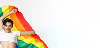 Ekin bluzlu ve kot pantolonlu mutlu eşcinsel kişinin dikey görüntüsü gökkuşağı bayrağını sallıyor, lgbtq bayramını kutluyor, beyaz arka planda duruyor..