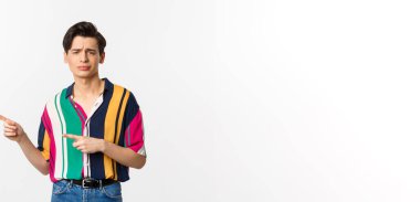 Şüpheci, küçümseyici bakışları olan ve sol elini gösteren, onaylamayan ve sevmeyen, beyaz arka planda duran eşcinsel bir adam..