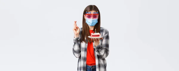 コロナウイルスの発生 ライフスタイルの社会的距離とお祝いの概念 眼鏡や医療マスクの熱狂的な幸せな女の子は 隔離で誕生日を祝います クロス指 ケーキで願いを作ります — ストック写真