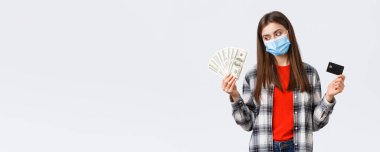 Para transferi, yatırım, covid-19 salgını ve ev konseptinden çalışma. Sağlık maskeli mutlu genç bir kadın para kazanıyor, dolarlara bakıp gülümsüyor ve kredi kartını gösteriyor.