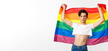 Kırpılmış bluzlu genç adam, yüzünde simle gururlu bayrağı kendine güvenle göndere çekiyor. lgbt bayrağıyla gülümseyen tuhaf insan..