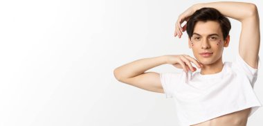 İnsanlar, lgbtq topluluğu ve yaşam tarzı konsepti. Dudak parlatıcısı ve göz altı ışıltısı olan küstah eşcinsel bir adamın yakın çekimi. Kameraya mutlu bir gülümsemeyle poz veriyor. Kırpılmış bluz giyiyor, beyaz arka plan..