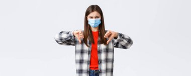 Coronavirus salgını, karantina, sosyal mesafeler ve duygu kavramı. Tıbbi maske takmış hayal kırıklığına uğramış ve kızgın genç bir kadın karşı oy kullandı, baş parmağını kaldırdı ve yüzünü buruşturdu.