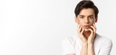 Millet, lgbtq ve güzellik konsepti. Yüzüne mavi ojeli parmaklarıyla dokunan, beyaz arka planda duran güzel eşcinsel adamın yakın çekimi..