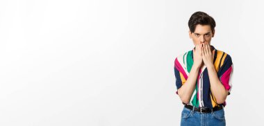 Meraklı görünen genç bir gay adamın görüntüsü, el ele tutuşmuş, dedikodu yapıyor, beyaz arka planda duruyor..