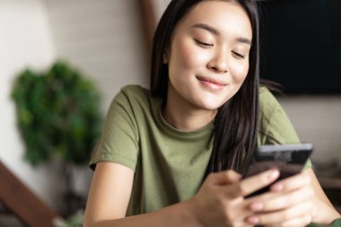 Gülümseyen Koreli kadın cep telefonu sohbet uygulamasına bakıyor rüya gibi yüzüyle, evinde koltukta oturuyor..